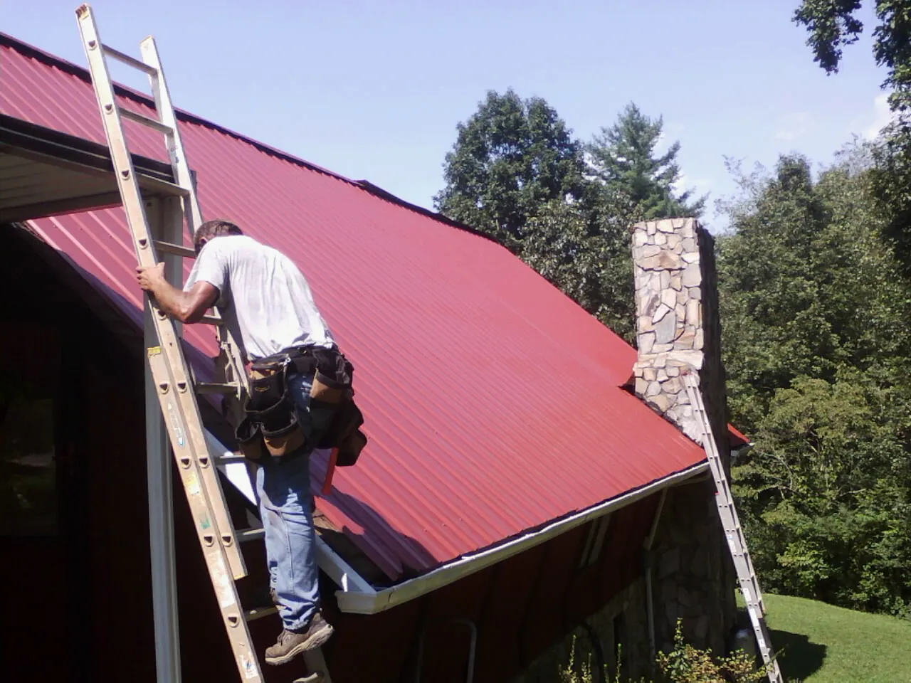 Roofer on a ladder beside a red metal roof and stone chimney — Soffit & Fascia Repair by Roofing Brothers in Sanbornville
